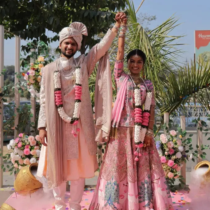 Couple in traditional attire with crochet floral garlands, standing outdoors with decorated background.